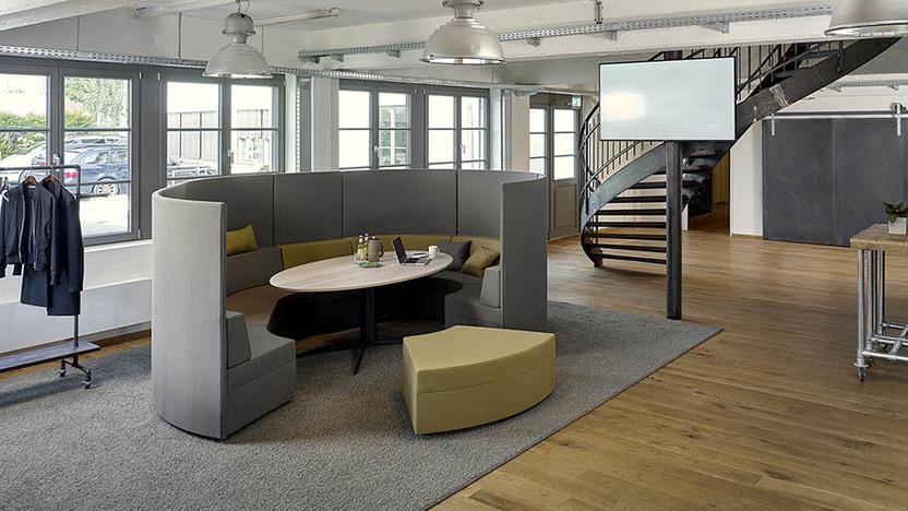 Runde Lounge um Rundtisch Grauer Loungebreich rund um einen Holztisch vor Treppe im Raum